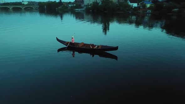 Aerial view of a strong man in hat rowing with the oar in a boat over the river background at night alt