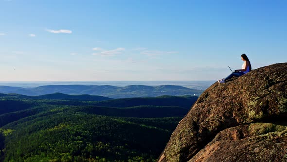 A Female Freelancer Works Sitting on the Edge of a Rocky Cliff in the Forest alt