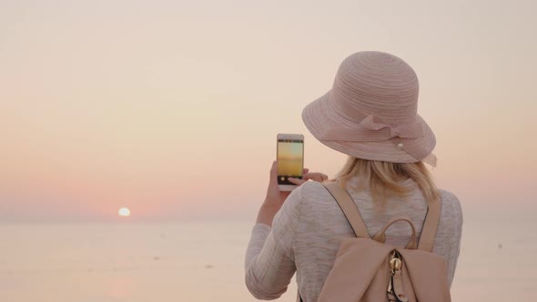 Young Stylish Girl in a Hat Takes Pictures of the Sea and a Pink Sunrise on a Smartphone alt