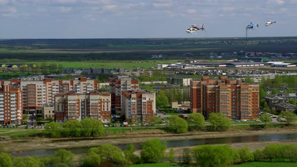 Three Helicopters Fly One After Other Over Brick Houses and Next to River on a Sunny Summer Day