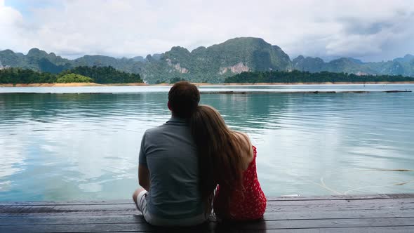 Happy Romantic Couple Sitting Together on Wooden Bridge on Green Mountains Lake alt