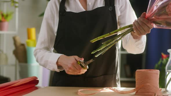Young Florist Cutting Off Ends of Flower Stems in Packaging Paper at Workplace alt