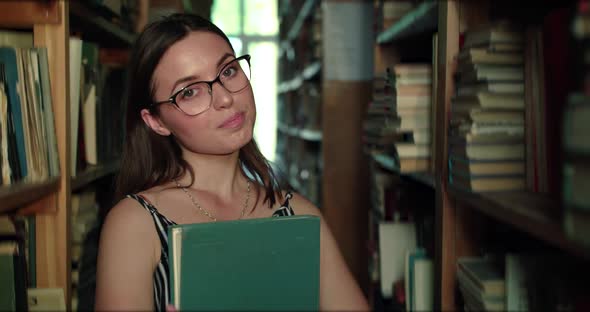 Girl in Glasses with Book in Hands Smiling Lightly at Camera in Library alt