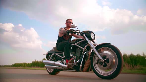 Stylish Biker with Tattoos Rides a Motorcycle on a Country Road at Sunset