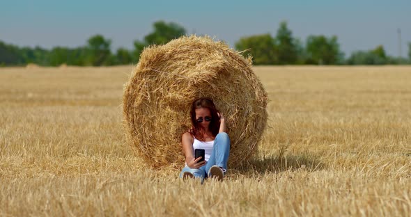 Young Woman Sits By a Large Golden Haystack in a Wheat Field and Takes a Selfie on Her Mobile Phone alt