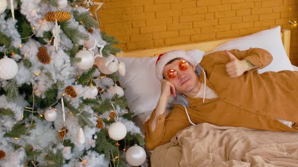 Young Man in Santa Hat and Fashionable Glasses Lying on Bed Near Christmas Tree alt