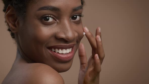 Attractive Black Lady Applying Cream Moisturizing Face Skin Beige Background alt