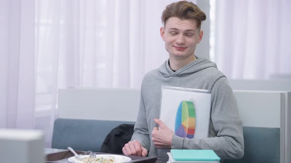Intelligent Millennial Young Male Student Posing with Diagram in Canteen Sitting at Table with Food alt