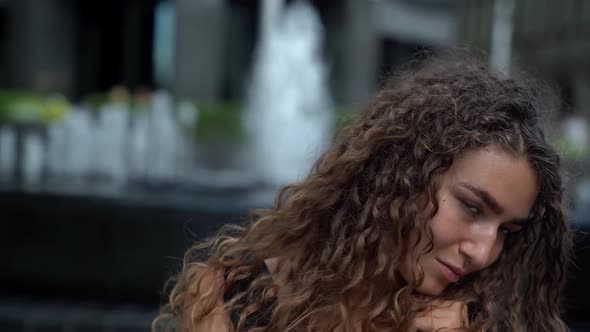 Portrait of a Young Woman with Curly Hair on the Background of a Fountain. Camera Zooms in alt