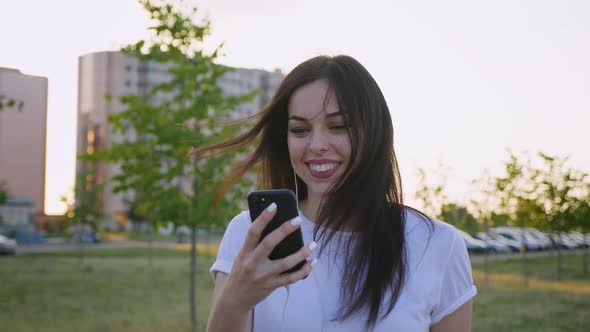 Portrait of Smiling Young Woman Using Smartphone While Walking on the City Street on Sunset