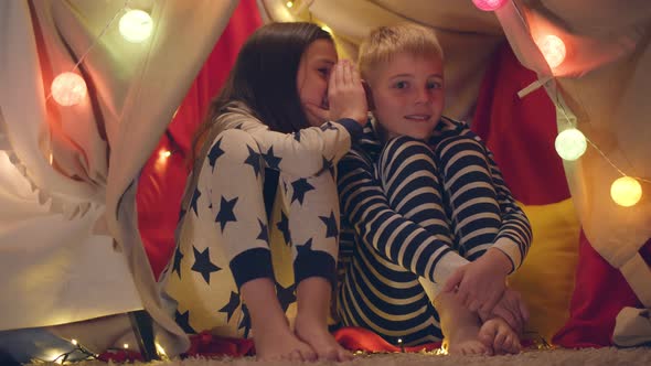 Happy Little Brother and Sister in Pajamas Telling Secrets Playing in Tent with Party Lights alt