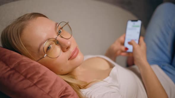 Woman in Glasses Looks at you Turns Away to Start Texting Using Mobile Phone Lying on Sofa alt
