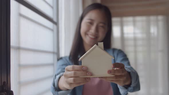 Asian woman freelance architect holding a model of home standing beside a window.