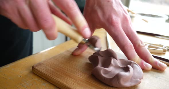 Close up on the hands of an artist carving and cutting brown modeling clay with a metal tool before alt