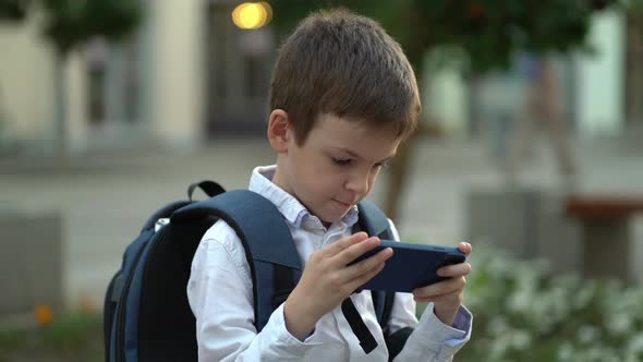 The Boy is Sitting on a Bench with a Backpack on His Back and Enjoying Time with the Phone
