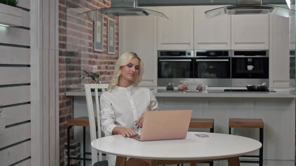 Blond Woman Freelancer in White Shirt Works at a Laptop at Home