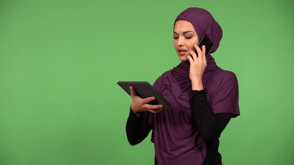 A Young Beautiful Muslim Woman Talks on a Smartphone and Holds a Tablet  Green Screen Background