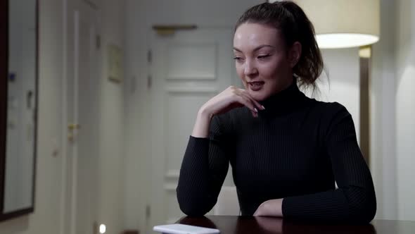 A Brunette Woman Sits at a Table in the Room and Picks Up Her Smartphone Smiling alt