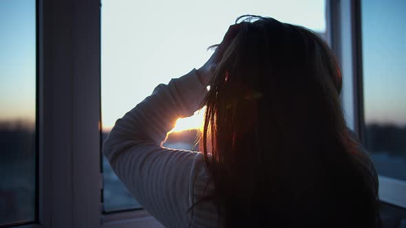 Woman Stands on the Balcony at Sunset and Looks Out the Window, Touches Her Hair alt
