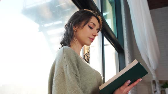 Beautiful Woman Sits Near Window and Reads a Book on Background of Sunset alt
