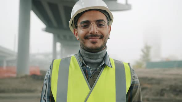 Portrait of Arab Man Wearing Helmet and Vest Smiling Standing Against Urban Construction Background alt