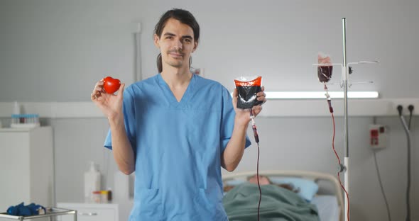 Male Nurse Holding Bag with Donor Blood and Red Rubber Heart Smiling at Camera alt