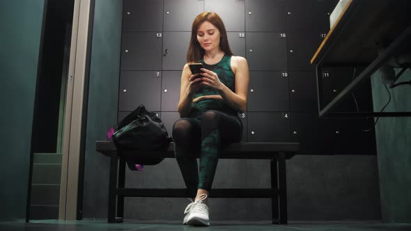 Young Woman in Sports Costume Sitting in Locker Room of Her Gym and Using Her Phone alt