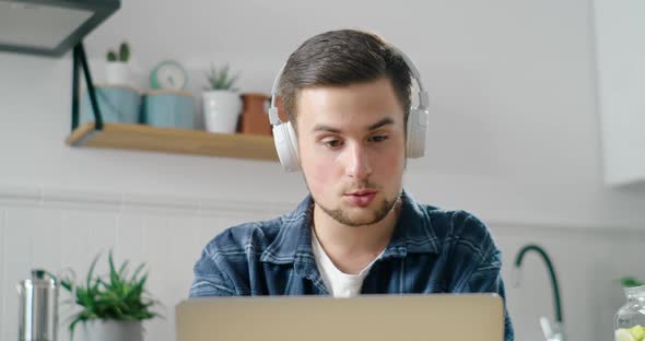 Young Man in Headphones Talks Online By Video Call on Laptop at Home in Kitchen