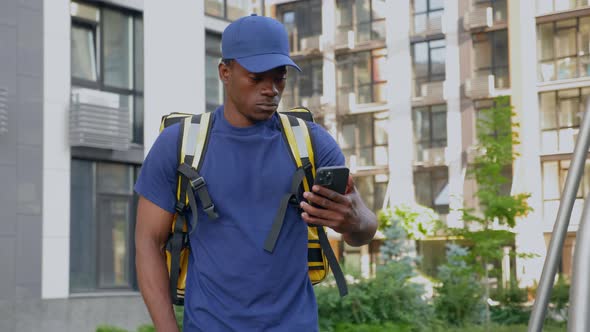 African Man Courier Delivery with Backpack Walks Street Watching in Smartphone alt