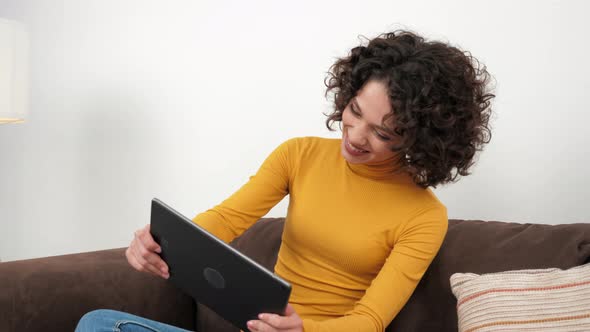 Curly Woman Emotionally Playing Video Game on Tablet Sitting on Couch at Home alt