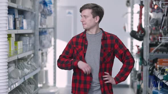 Uncertain Brunette Young Man with Brown Eyes Standing Between Rows in Hardware Store Thinking alt