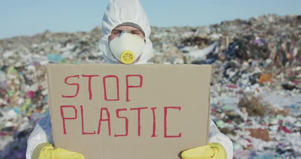 Man in Protective Suit Show Protest Sign "Stop Plastic" at Camera at Landfill alt