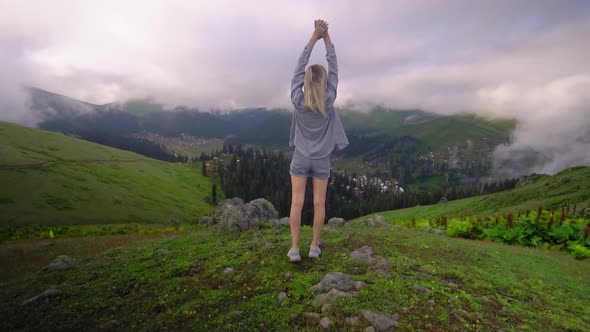 Young girl raised her hands up against background of clouds in mountains. alt