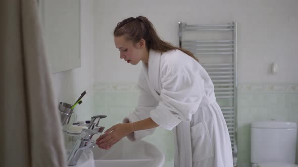 Slim Charming Caucasian Woman in White Bathrobe Turning on Faucet and Washing Face in Slow Motion alt