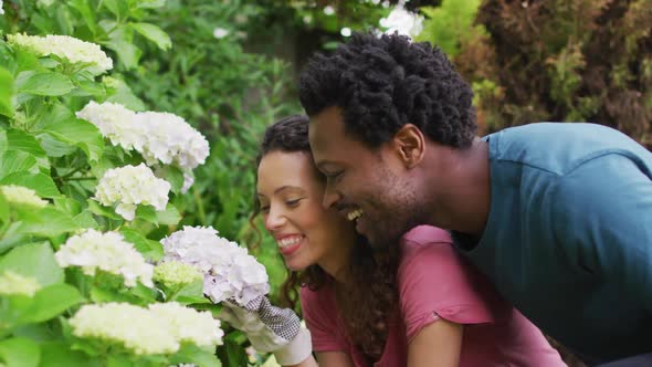 Happy biracial couple gardening together, smelling flowers and laughing alt