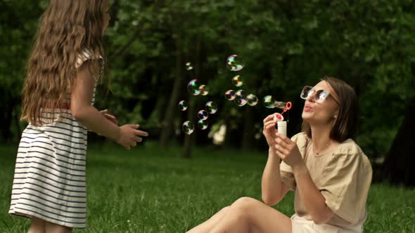Beautiful Young Woman Sits on Green Grass in the Park and Blows Soap Bubbles