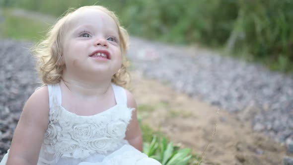 Little Funny Cute Blonde Girl Child Toddler with Curls in White Dress and with Mud on Her Face Sits
