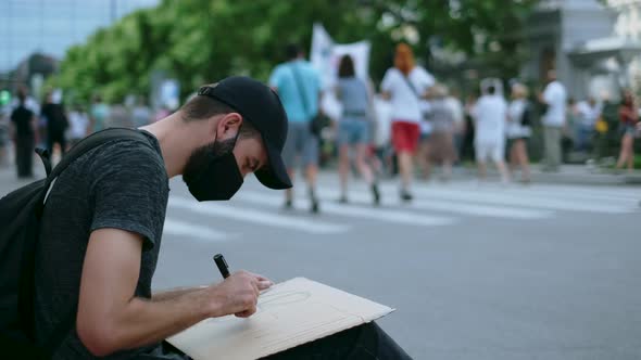 Sitting Demonstrator Rebel in Mask Draws Banner Placard Poster Sign with Marker alt