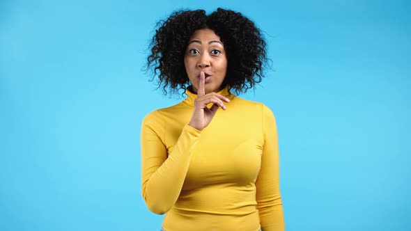Smiling African Woman with Curly Hair Holding Finger on Her Lips Over Blue Background alt