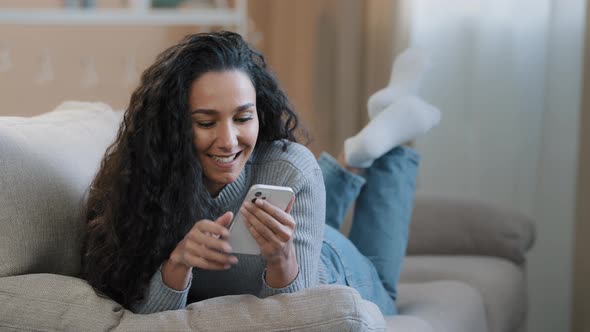 Happy Hispanic Young Woman Lying on Sofa Typing Message on Phone Smiling Carefree Girl Communicate alt