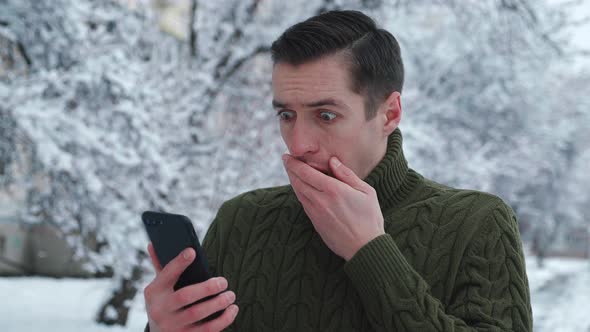 Shocked Man Reacting Message on Smartphone Covers His Mouth in Fright While Standing Winter Snowy alt