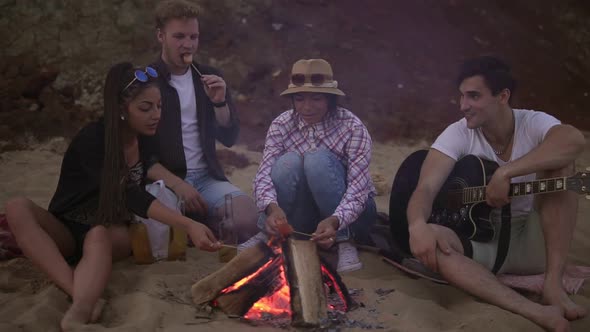 Group of Young and Cheerful People Sitting By the Fire on the Beach in the Evening Grilling Sausages alt