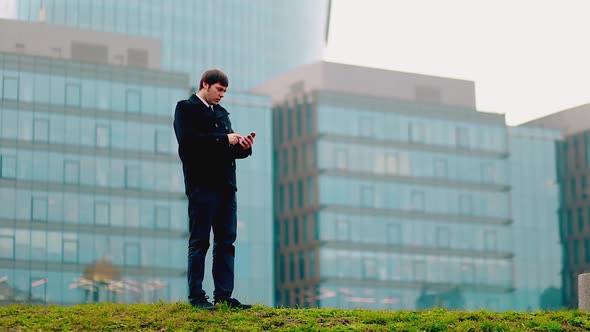 A Man Dials a Number on the Phone Against the Backdrop of Highrise Buildings