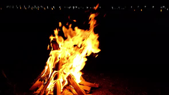 Bonfire lit at night with the lights of the night city reflected in the lake alt