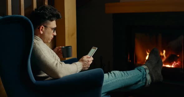 Man Reads eBook on Electronic Reader By Fireplace in Cozy Room at Night