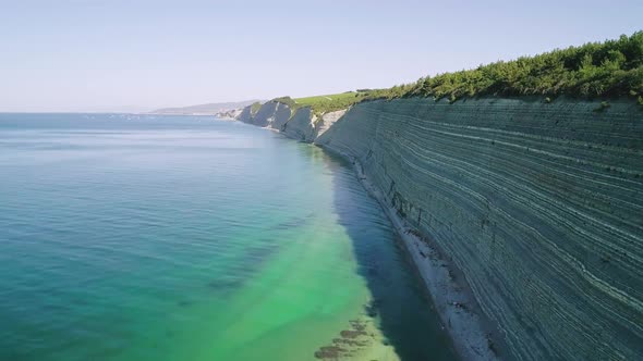 Aerial of High Steep Cliffs Covered with Pine Trees and Wild Pebble Beach with Turquoise Sea Water alt