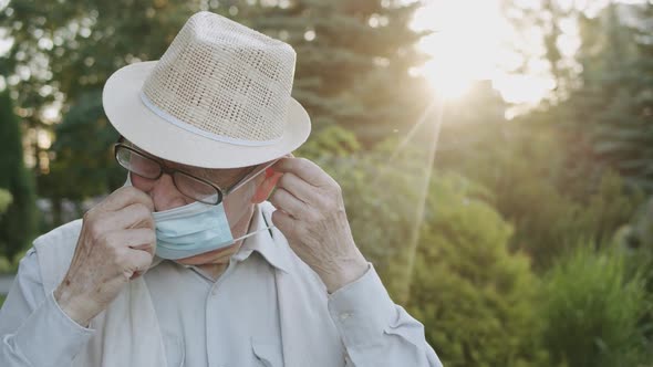 Senior Man with Glasses Wears Medical Mask and Looking Into Camera Outdoors alt