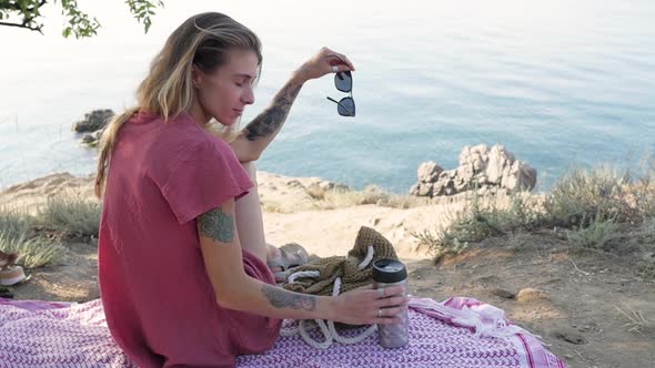 Young Woman Sitting on a Blanket on the Beach By the Sea Looking in Front of Her and Holding a alt