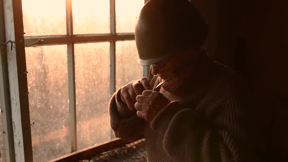 Elderly Man with Glasses Picks Up Military Helmet in Dust and Putting Helmet on Head in House Window alt