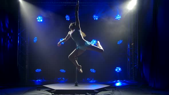 A Cute Young Woman Spins on a Pole in Slow Motion. Blue Background in the Studio. Sports Pole alt